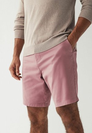 Shorts en coton rose avec poches latérales, coupe ajustée et devant plat. Portés avec un pull beige, présentant un look décontracté.