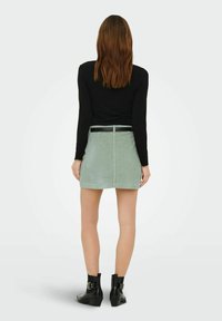 Femme de dos portant un haut à manches longues noir, une jupe en velours côtelé vert clair avec une ceinture, et des bottines noires à boucles.