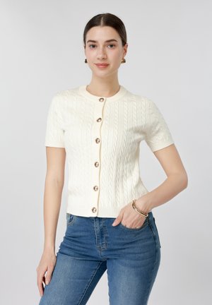 Nő krémszínű, rövid ujjú, fonott mintás kardigánban gyöngygombokkal és kék farmerral, egyik kezét zsebében tartva, enyhe sminkkel pózol.