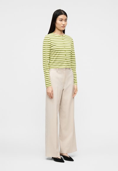 Γυν αίκα που στέκεται φορώντας ένα πράσινο και λευκό ριγωτό ζακέτα με κουμπιά, μπεζ παντελόνι με φαρδιές παντελόνες και μαύρες μυτερές γόβες σε μονόχρωμο φόντο.