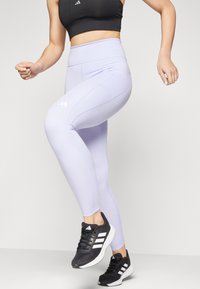 Světle fialové legíny s vysokým pasem a hladkou texturou, s černým kontrastním prošíváním a logem Adidas. Spárované s černými teniskami.