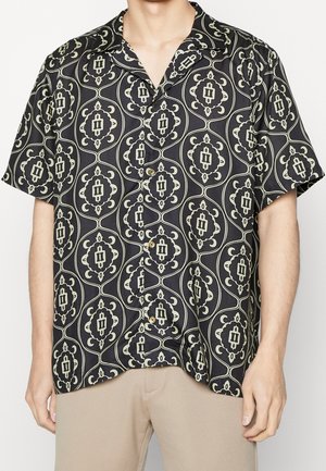 Camicia nera a maniche corte con bottoni e motivo floreale geometrico beige, indossata con pantaloni beige, vista dal collo ai fianchi.