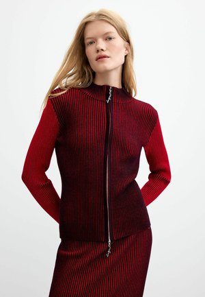 Femme aux longs cheveux blonds portant un pull zippé rouge et noir à rayures près du corps et une jupe assortie sur fond blanc uni.