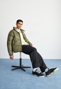 Olīvu zaļš pūkains žakets, krēmkrāsas T-krekls, melnas platās bikses, melnas kedas ar baltām zolēm un dzelteniem akcentiem, sēž uz griežamā krēsla.