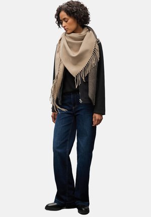 Écharpe en laine beige avec des franges, portée sur un haut noir et un vêtement d'extérieur gris, associée à un jean bleu foncé et des bottines noires.