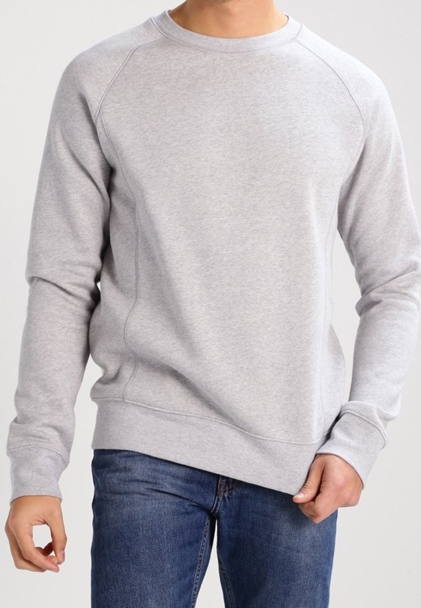 Uomo che indossa un pullover grigio chiaro a girocollo e jeans blu, che tiene l'orlo del pullover con la mano destra.