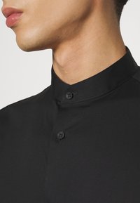 Camicia nera con colletto alto, dotata di due bottoni neri. Il tessuto è liscio con una leggera lucentezza, enfatizzando un design pulito e minimalista.