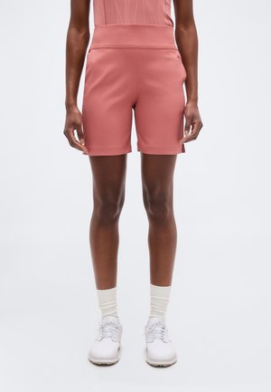 Osoba ubrana w pudrowo różowe krótkie spodenki sięgające do kolan, pasujący top, białe skarpetki i białe buty na sznurowadła stojąca na tle jednolitym.