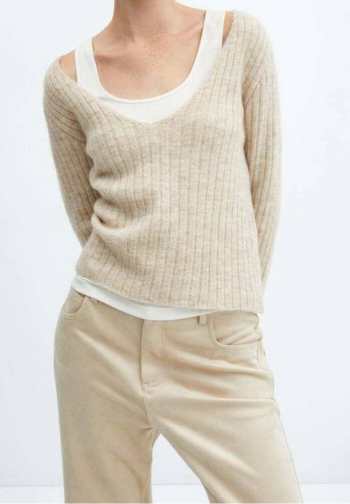 Femme portant un pull beige côtelé à col en V sur un débardeur blanc, associé à un pantalon beige, debout devant un fond blanc uni.