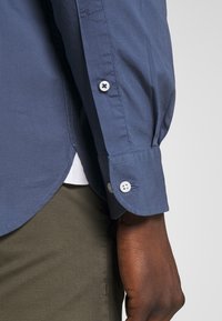 Camicia button-down in cotone blu scuro con maniche lunghe. Presenta bottoni bianchi e un orlo curvo, indossata con pantaloni verde oliva.