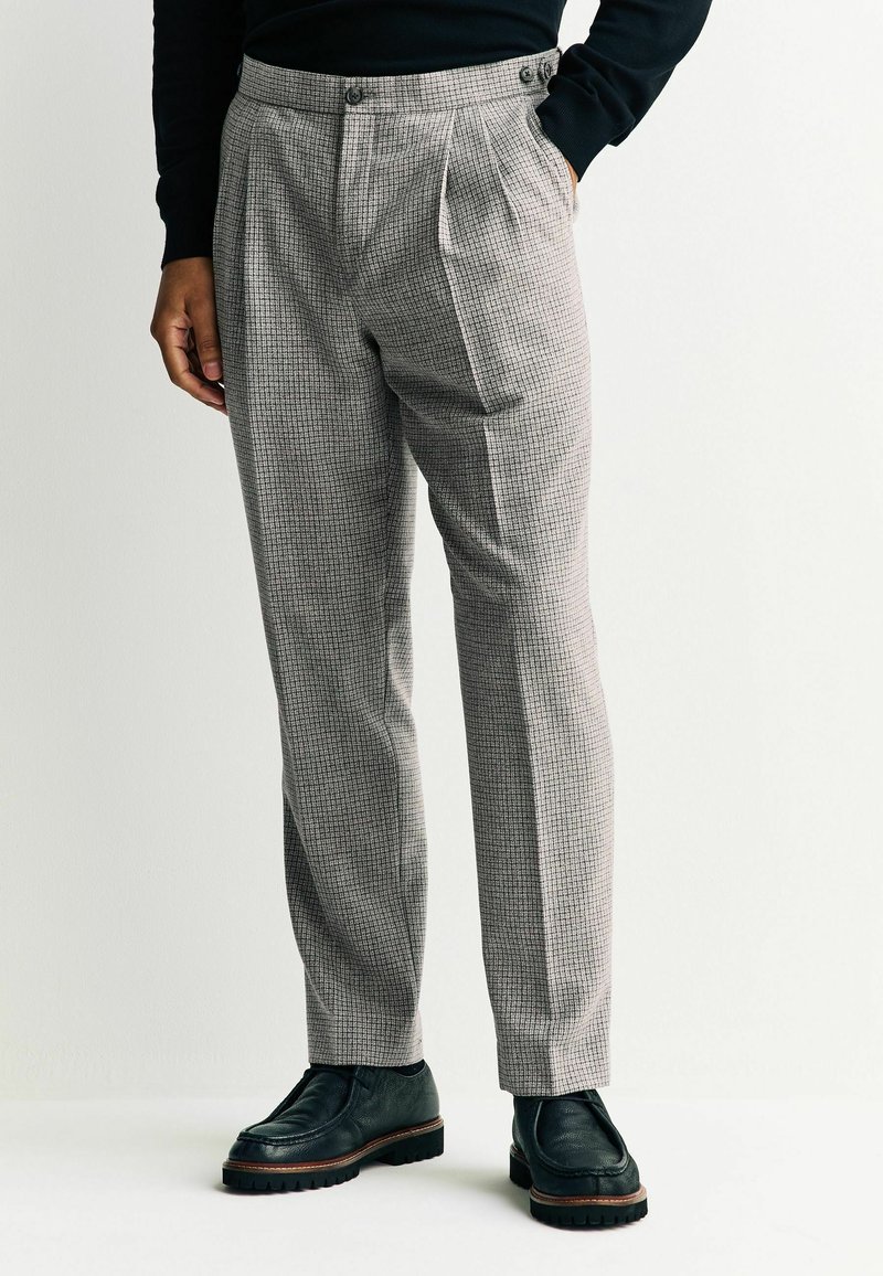 Pantalon gris à carreaux avec un devant plissé, taille haute et poches latérales, assorti à des chaussures en cuir noir avec une semelle épaisse.