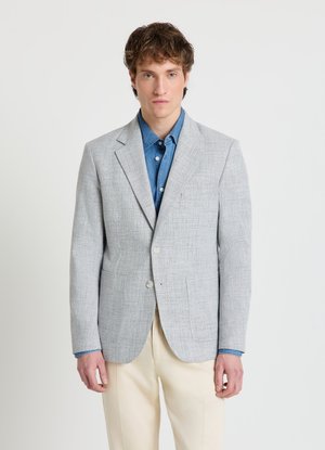 Homme portant un blazer gris clair sur une chemise bleue boutonnée et un pantalon crème, debout devant un fond blanc uni.