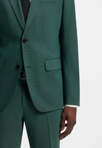 Veste et pantalon de costume sur mesure vert foncé portés avec une chemise blanche et une cravate rayée noire, montrés du milieu du torse à la main.