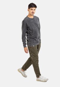 Man walking, wearing a washed black long-sleeve shirt, olive green cargo pants, and white sneakers on a white background.