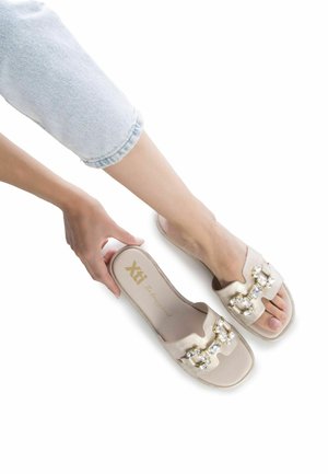 Chanclas de color beige con adornos de joyas, diseño de punta cuadrada, material suave y suela plana, presentadas sobre un fondo blanco.