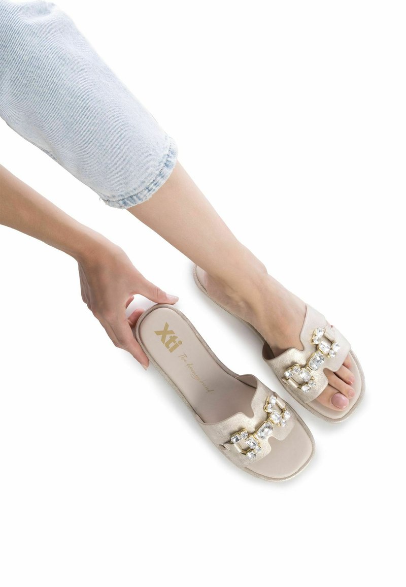 Chanclas de color beige con adornos de joyas, diseño de punta cuadrada, material suave y suela plana, presentadas sobre un fondo blanco.