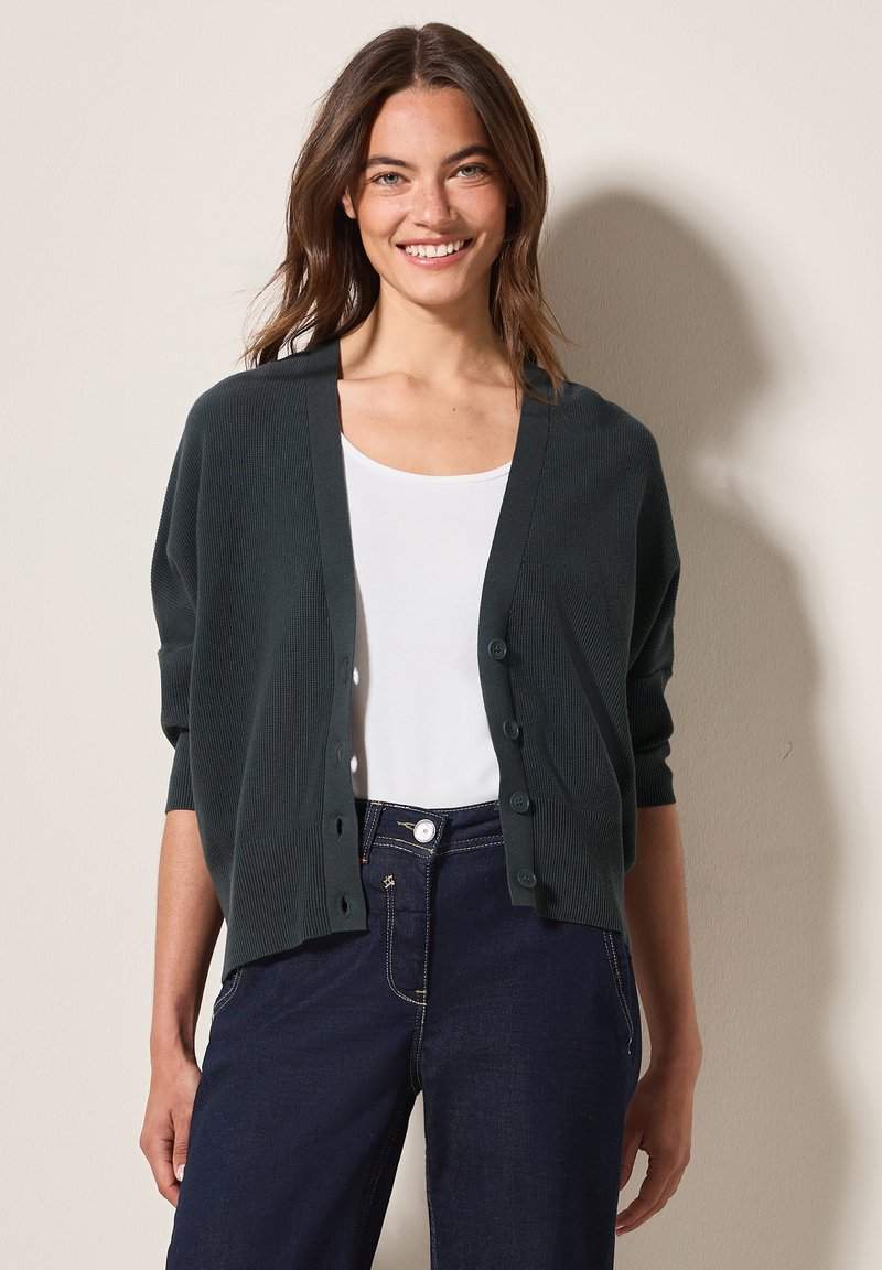 Donkergroen gebreide cardigan met een V-hals, knoopsluiting aan de voorkant en driekwartmouwen, gecombineerd met een witte blouse en donkere spijkerbroeken.