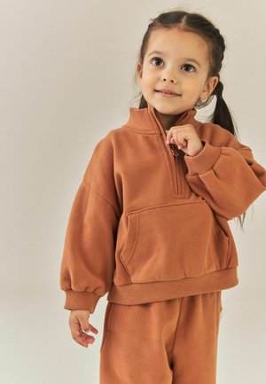 Ung pige med mørkt hår sat op i to hestehaler iført en brun lynlåstrøje og matchende bukser, der ser let opad med et blidt smil.