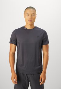 Mand med kort hår iført en mørkegrå t-shirt med korte ærmer og matchende shorts foran en ensfarvet lys baggrund.