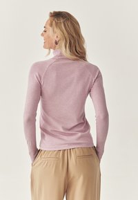 Donna con capelli biondi, che indossa un maglione a collo alto rosa chiaro e pantaloni beige, in piedi con le mani nelle tasche, voltata di spalle.