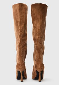 Brun suede ylipolvensaappaat, joissa on kapea stiletto-korko, kuvattuna takaa yksinkertaista valkoista taustaa vasten.