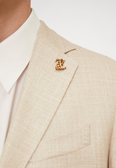 Giacca di suit beige con texture e un leggero peso, caratterizzata da una spilla per bavero in tonalità oro con design in lettere corsive. Sotto, una camicia bianca fresca.