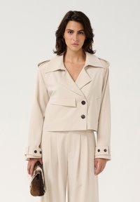 Beige cropped Jacke mit großen Revers, zwei vorderen Knöpfen und Tasche. Die langen Ärmel haben geknöpfte Manschetten. Begleitet von einer strukturierten Clutch.