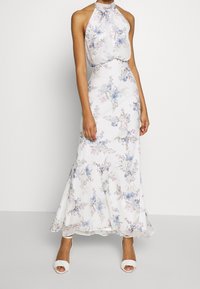 Kvinna som bär en ärmlös, höghalss, vit maxiklänning med blå och lila blommönster och vita klacksandaler.