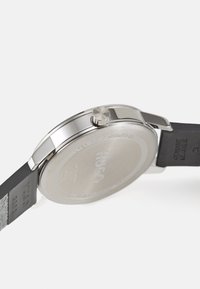 Edelstahl-Gehäuseboden mit eingeprägtem Markennamen. Schwarzes Gummiband mit strukturierten Akzenten. Wasserdichtheit angezeigt.
