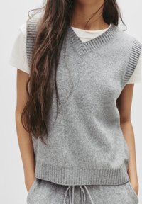 Grauer, gestrickter ärmelloser Pullover mit V-Ausschnitt und gerippten Details, über einem weißen T-Shirt getragen. Weicher Stoff, lässige Passform.