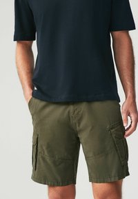 Pantaloni scurți cargo de culoare verde măsliniu, cu multiple buzunare laterale, confecționați dintr-un material ușor. Asortați cu un tricou negru cu mânecă scurtă.