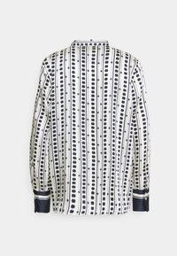 Chemise blanche à manches longues avec des motifs verticaux de lignes et de points noirs, poignets noirs à rayures blanches, vue de dos.