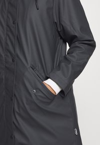 Svart vattentät kappa med en slät yta, tryckknappar, sidofickor och justerbar huva. Vitt manschettdetalj synligt vid ärmen.