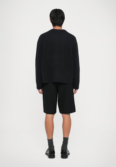 Uomo che indossa un maglione di lana nero, pantaloni corti neri fino al ginocchio, calze grigie scure e scarpe nere, in piedi di fronte a uno sfondo bianco.