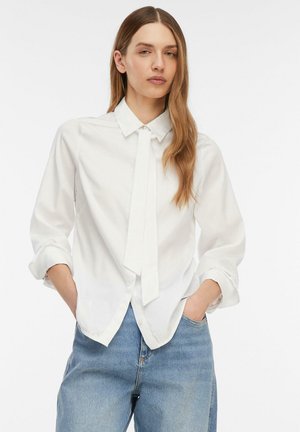 Frau mit langen Haaren, die ein weißes Hemd mit Knöpfen und Krawattendetail sowie hellblaue Jeans trägt und mit den Händen in den Taschen vor einem schlichten Hintergrund steht.