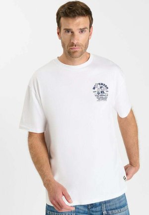 Man die een wit T-shirt met korte mouwen draagt met blauwe grafische tekst aan de linkerkant van de voorkant en lichtblauwe spijkerbroek, staand tegen een effen witte achtergrond.