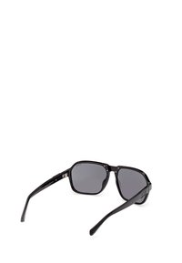 Lunettes de soleil carrées à monture noire avec des verres teintés foncés, présentées en angle avec les branches déployées sur un fond blanc.