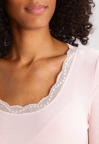 Vaaleanpunainen ribbitekniikalla valmistettu paita, jossa on herkät pitsikoristeet kaula-aukossa, sileä pinta ja lyhyet hihat.