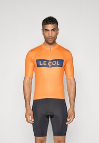Krátký rukáv cyklistický dres v jasně oranžové barvě s modro-námořnickým logem "LE COL" na prsou, doplněný o těsně padnoucí černé kraťasy.