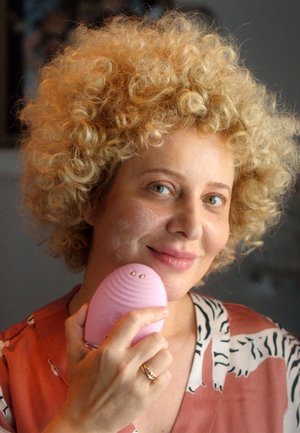 Pink silicone facial cleansing device with ridged texture held against cheek, featuring two gold accents. User has curly blonde hair.