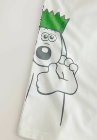 Tricou alb cu un personaj de desen animat care are o coroană verde, ochi mari, o gură deschisă și un gest de aprobă cu degetul mare. Material din bumbac moale.