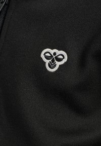 Tessuto nero con finitura testurizzata presenta un logo di un'ape ricamato in bianco. Il design include un fiocco e forme geometriche.