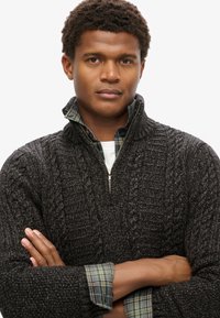 Dunkelgrauer Strickpullover mit hohem Kragen und Reißverschluss. Darunter ein grün kariertes Hemd mit sichtbarem Manschette.