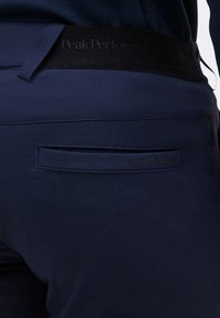 Gros plan sur la poche arrière et la ceinture d'un pantalon bleu marine avec le logo "Peak Performance" embossé.