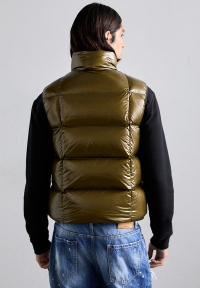 Olive green puffer vest with high collar, shiny texture, and quilted design. Worn over a black long-sleeve shirt and paired with blue distressed jeans.