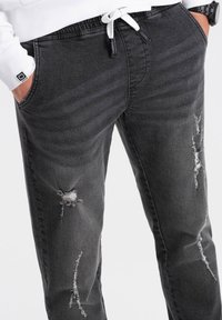 Pantalones jogger de mezclilla gris con cintura ajustable, que presentan detalles desgastados y múltiples bolsillos para un look informal.
