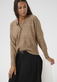 Pull en tricot marron clair avec un col en V, des manches longues et un ourlet côtelé, associé à une jupe noire avec une ceinture en métal argenté.