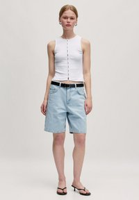 Weißes ärmelloses Crop-Top mit einer Druckknopfverschluss vorne, kombiniert mit hellblauen Denim-Bermuda-Shorts und einem schwarzen Gürtel. Gestylt mit schwarzen Sandalen.