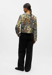 Veste courte à motif floral en tissu multicolore avec des manches longues, associée à un pantalon large en velours côtelé noir et des chaussures noires.