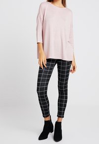 Frau trägt ein hellrosa, lockeres Oberteil, schwarz-weiße karierte Slim Pants und schwarze Stiefeletten vor einem einfarbigen Hintergrund.